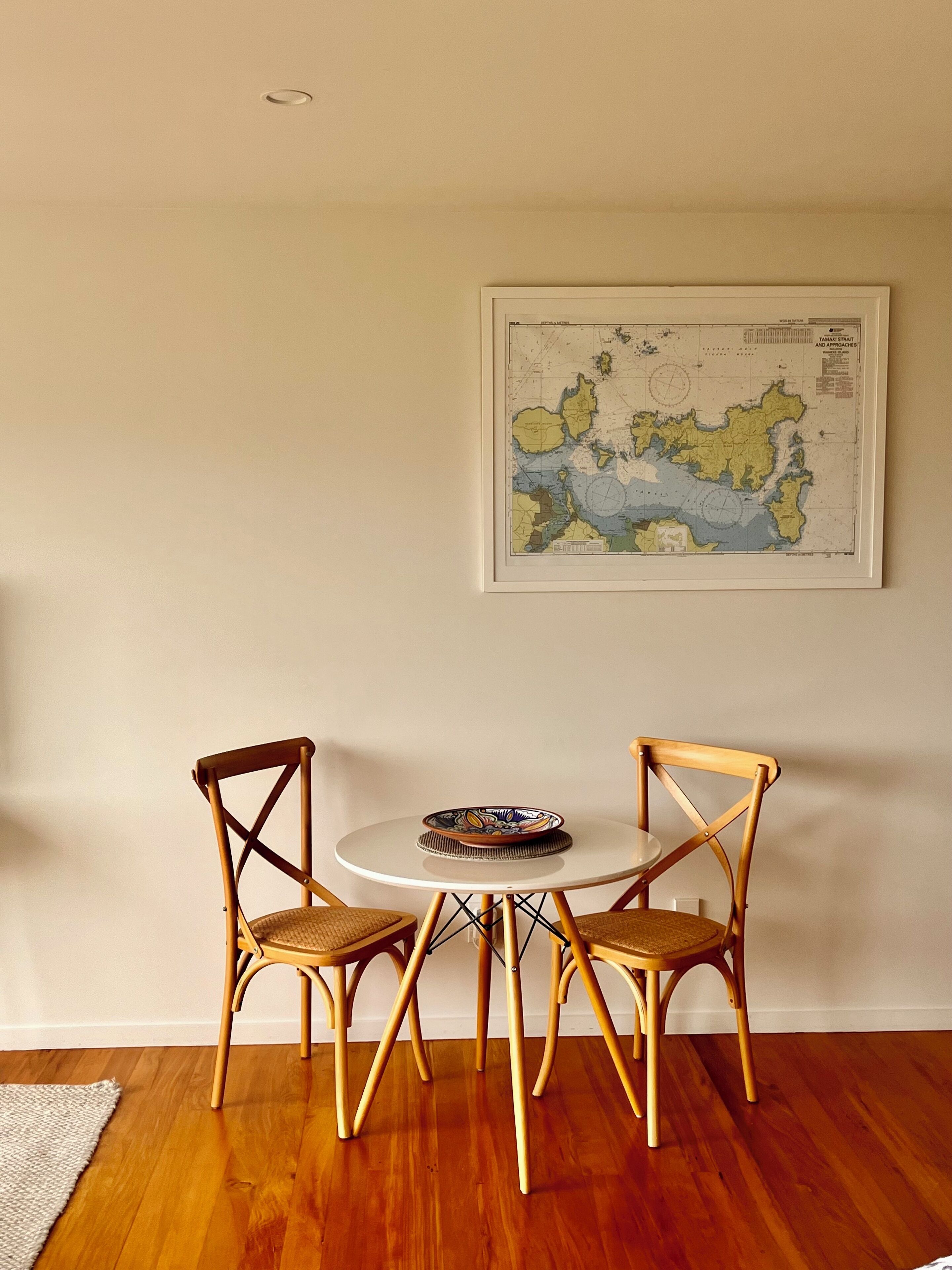 A dining area with a round table and two wooden chairs, a framed map on the wall, and a wooden floor.