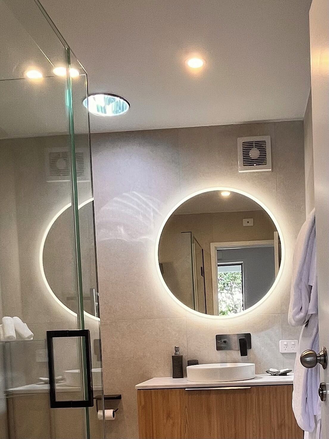 Un baño moderno con cabina de ducha de vidrio, un espejo redondo y un mueble de tocador de madera.