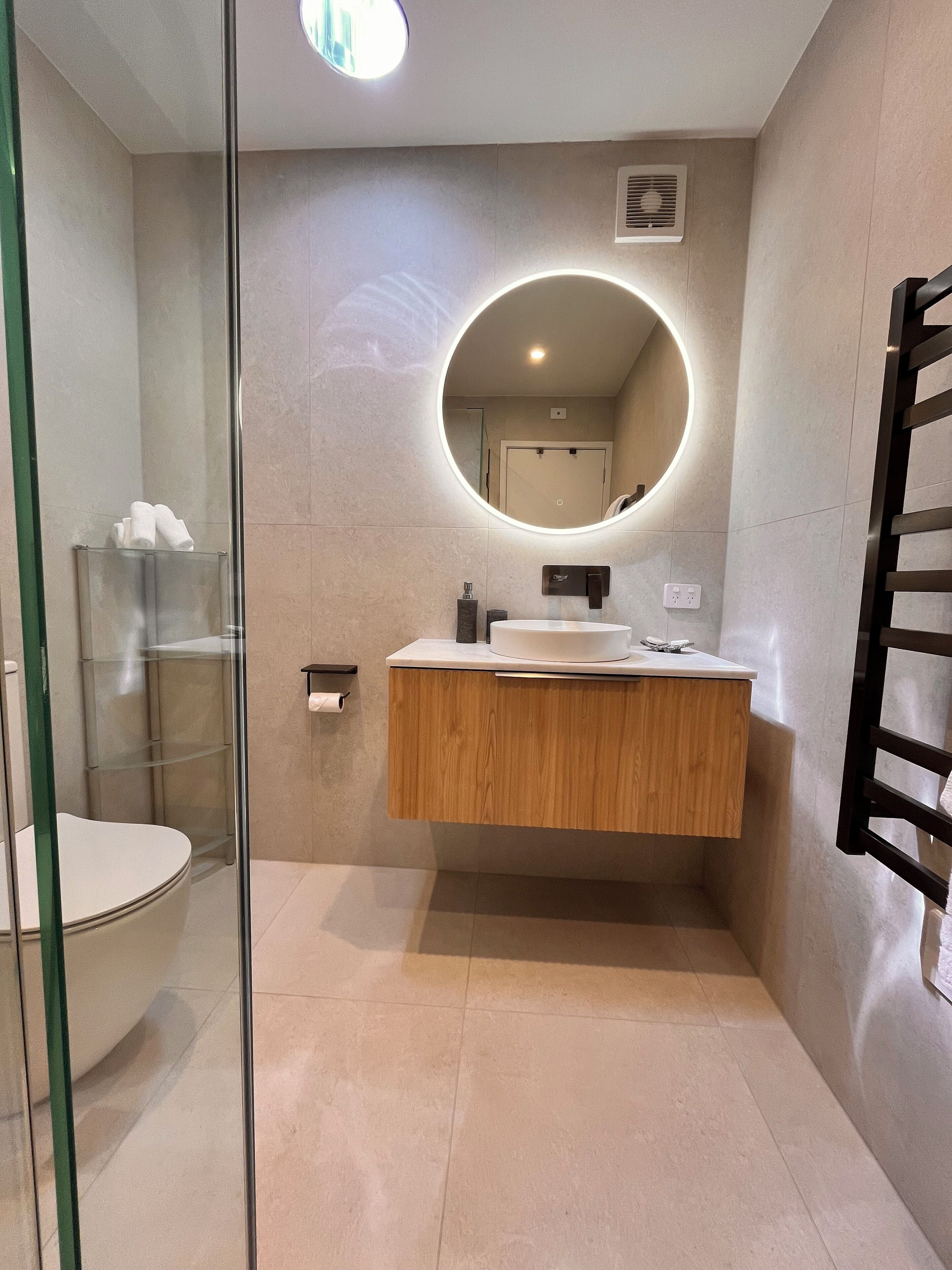 Une salle de bain moderne avec une douche à l’italienne en verre, un meuble-lavabo en bois avec un miroir rond et des toilettes blanches.