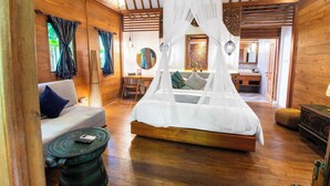 A wooden-floored bedroom with a canopy bed, a sofa, a small round table, and a chair. - Natya Hotel Tanah Lot (Tabanan)