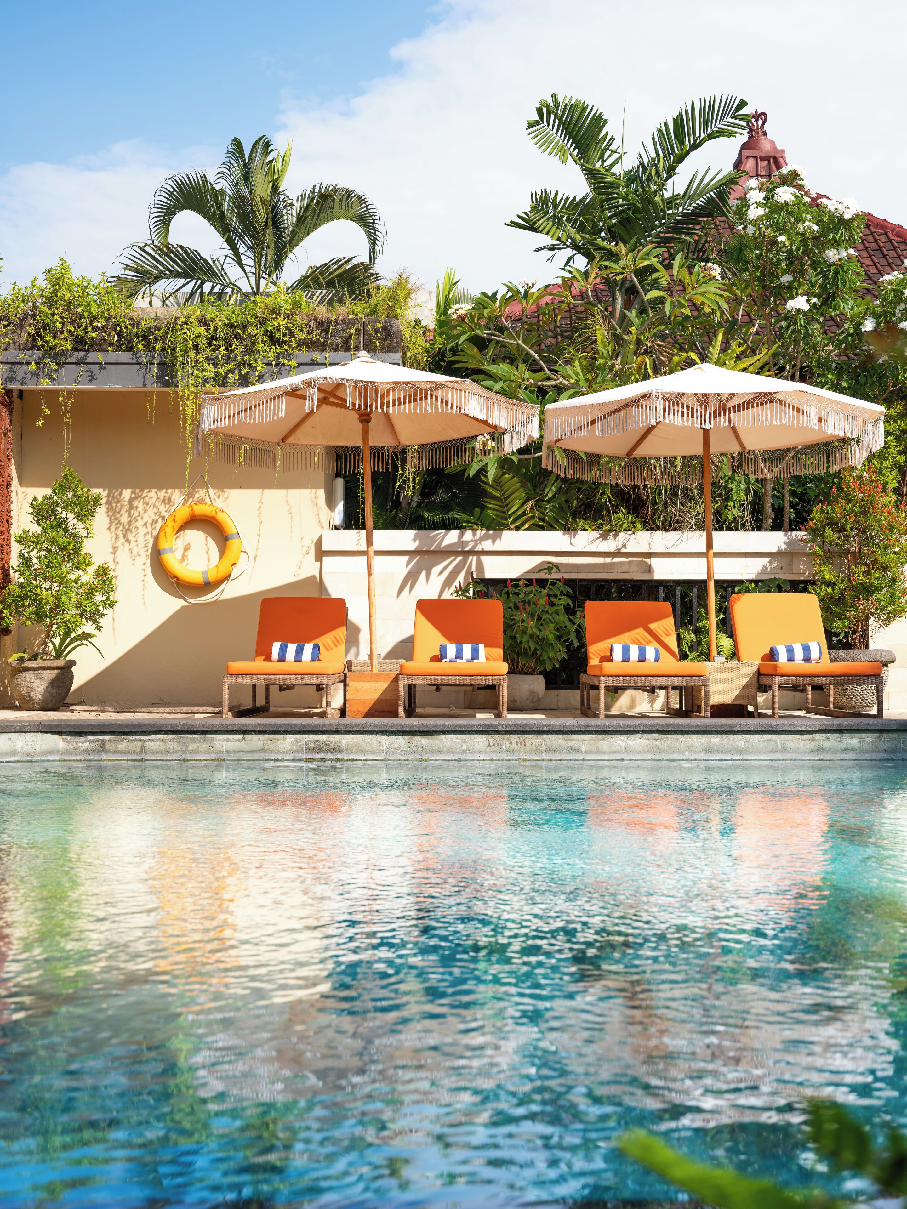 A poolside area with lounge chairs and umbrellas, a lifebuoy, and lush greenery in the background.
