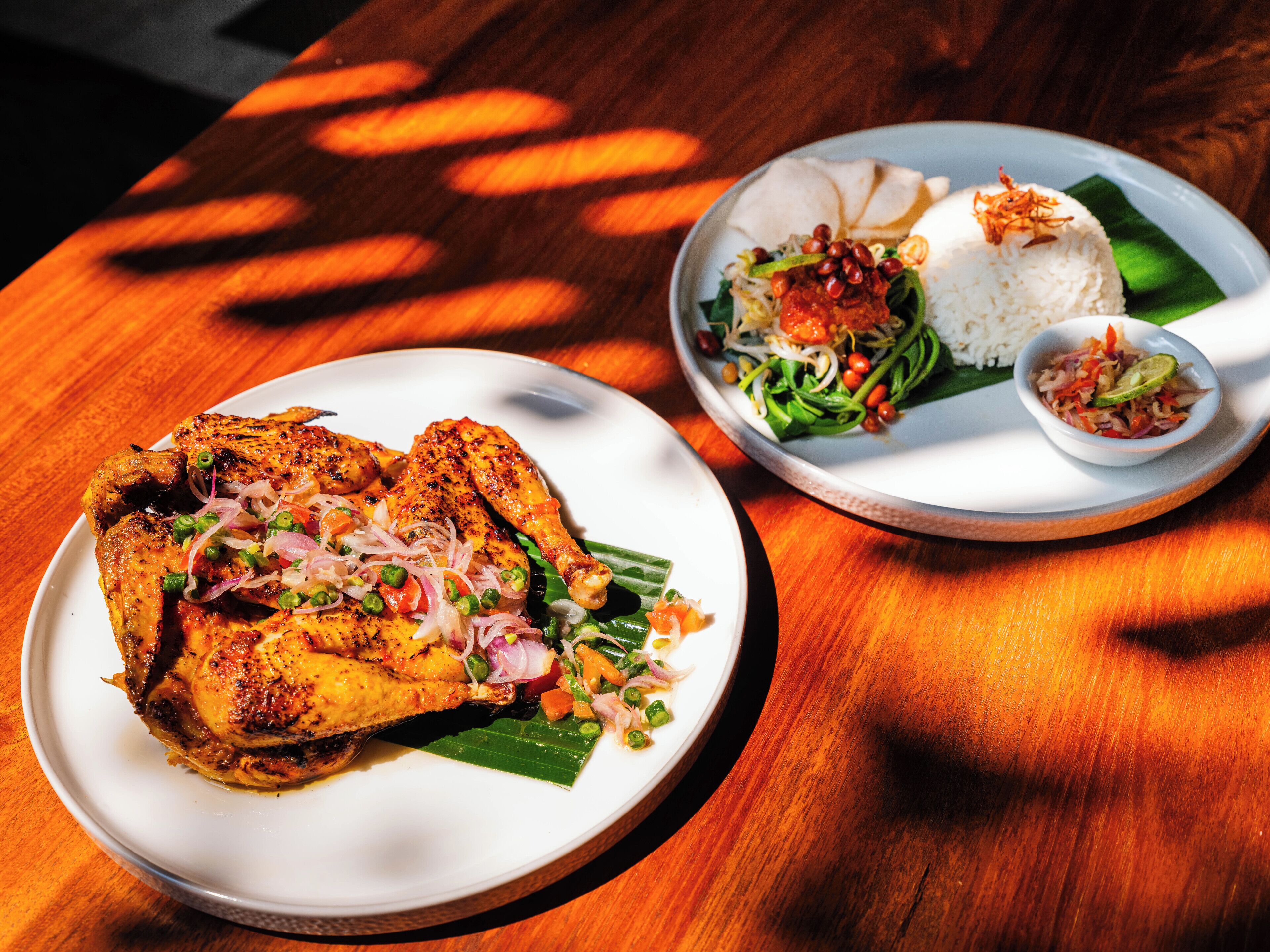 A wooden table with two plates of food, one with grilled chicken and the other with rice and vegetables.