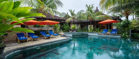 A swimming pool with blue lounge chairs and orange umbrellas, surrounded by palm trees and a traditional building.