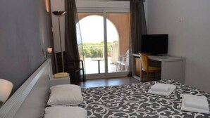 A hotel room with a bed, a desk with a television, a chair, and a view of a balcony with outdoor furniture. - Hotel Orizonte (Cervione)