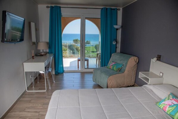 A hotel room with a bed, a desk, a chair, and a balcony with a view of the sea.