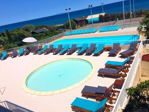 A pool area with sun loungers and a central swimming pool, surrounded by a fence and overlooking the ocean.
