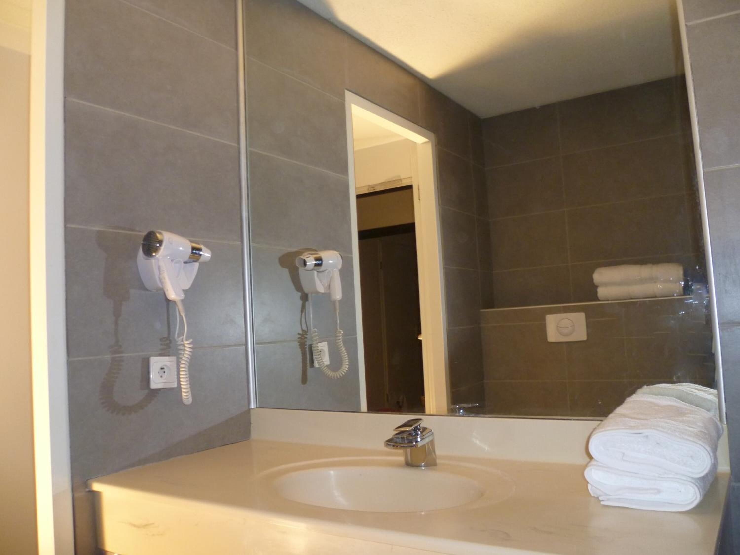 Ein Badezimmer mit Waschbecken, zwei an der Wand montierten Haartrocknern, einem Spiegel und einem Handtuchhalter mit gefalteten Handtüchern.