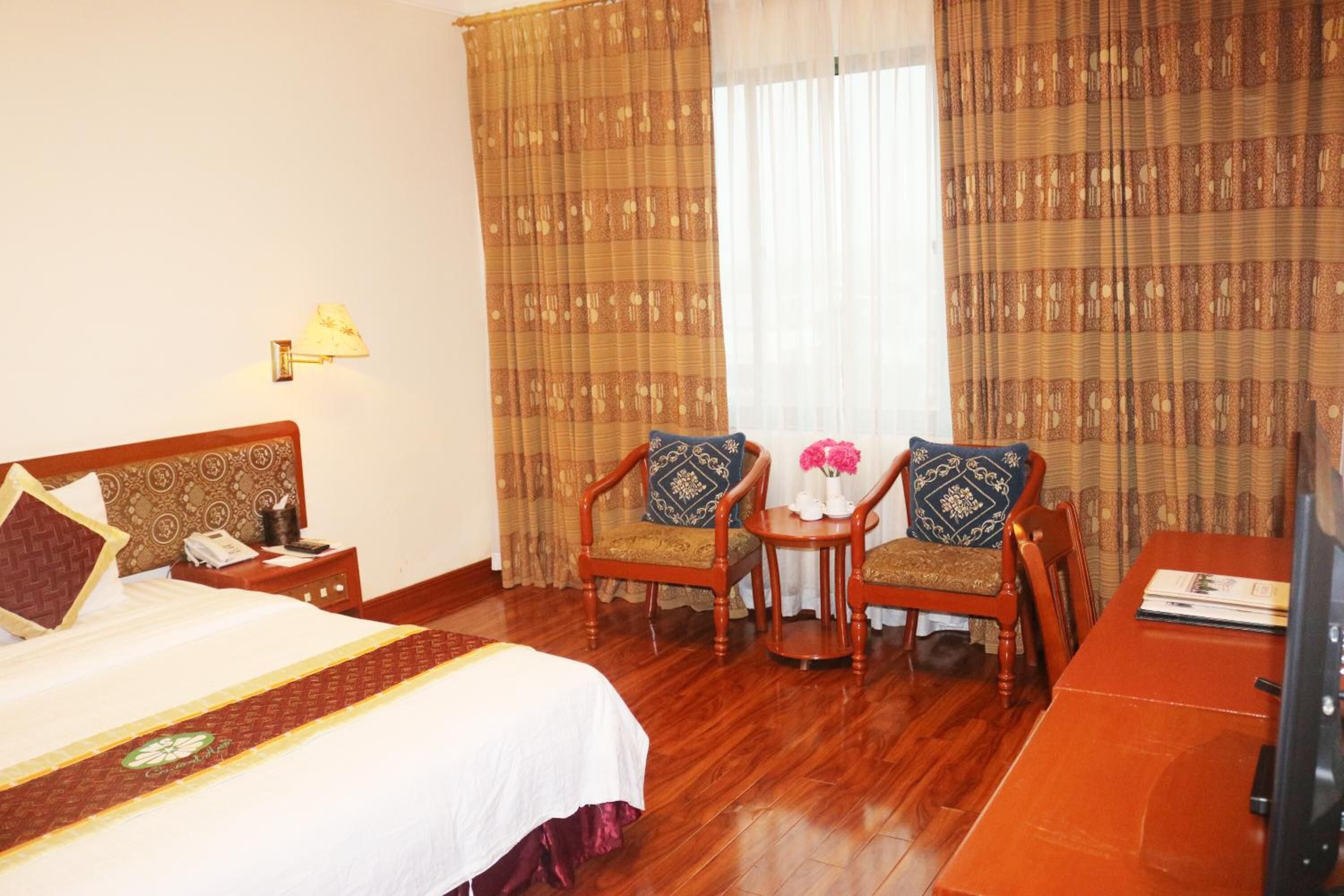 A hotel room with a bed, two wooden chairs, a small table with flowers, a desk with a computer, and curtains.
