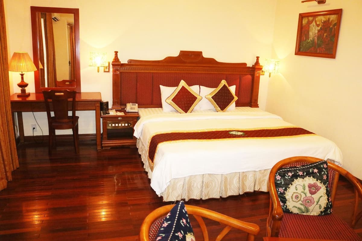 Une chambre d’hôtel avec un lit en bois, un bureau avec une lampe, une chaise avec un coussin à motifs floraux et un tableau au mur.
