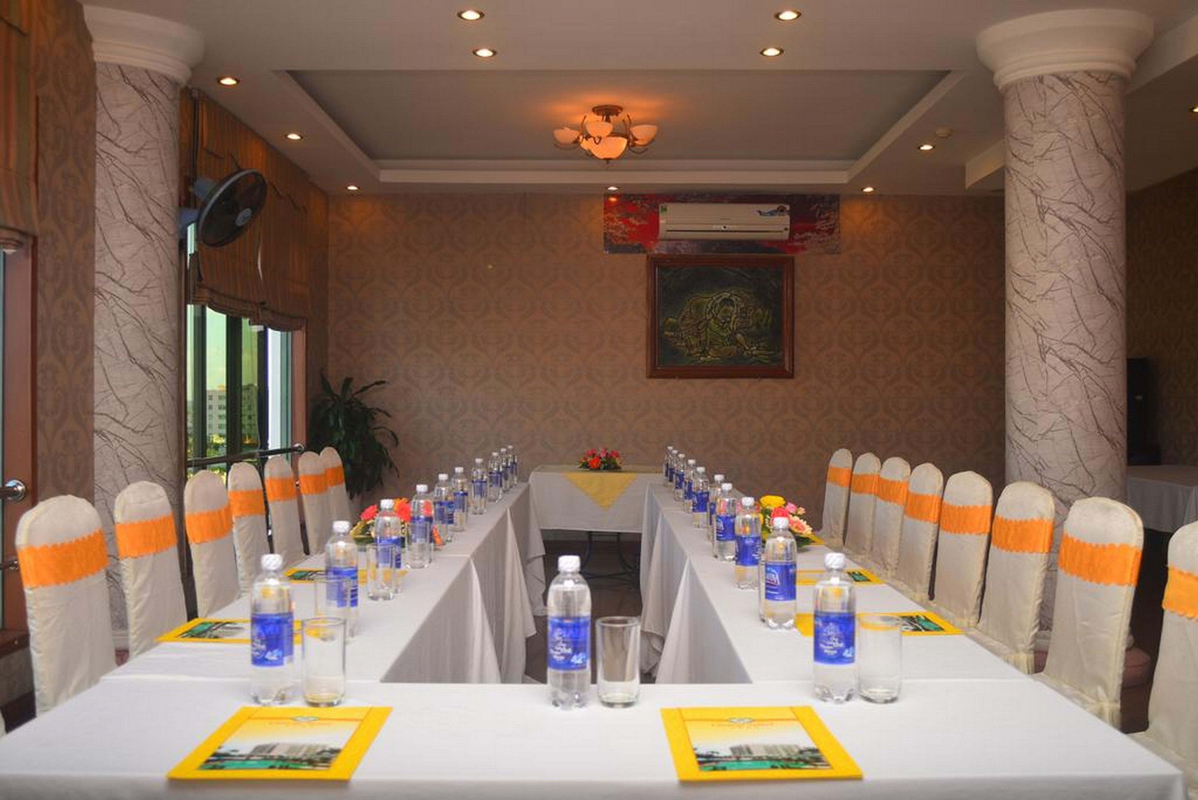 A conference room with a long table, chairs with orange sashes, water bottles, and a ceiling fan.