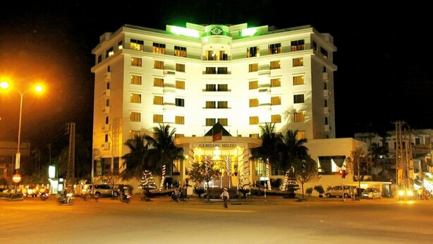 Um hotel com vários andares e entrada central, rodeado por palmeiras e postes de luz.