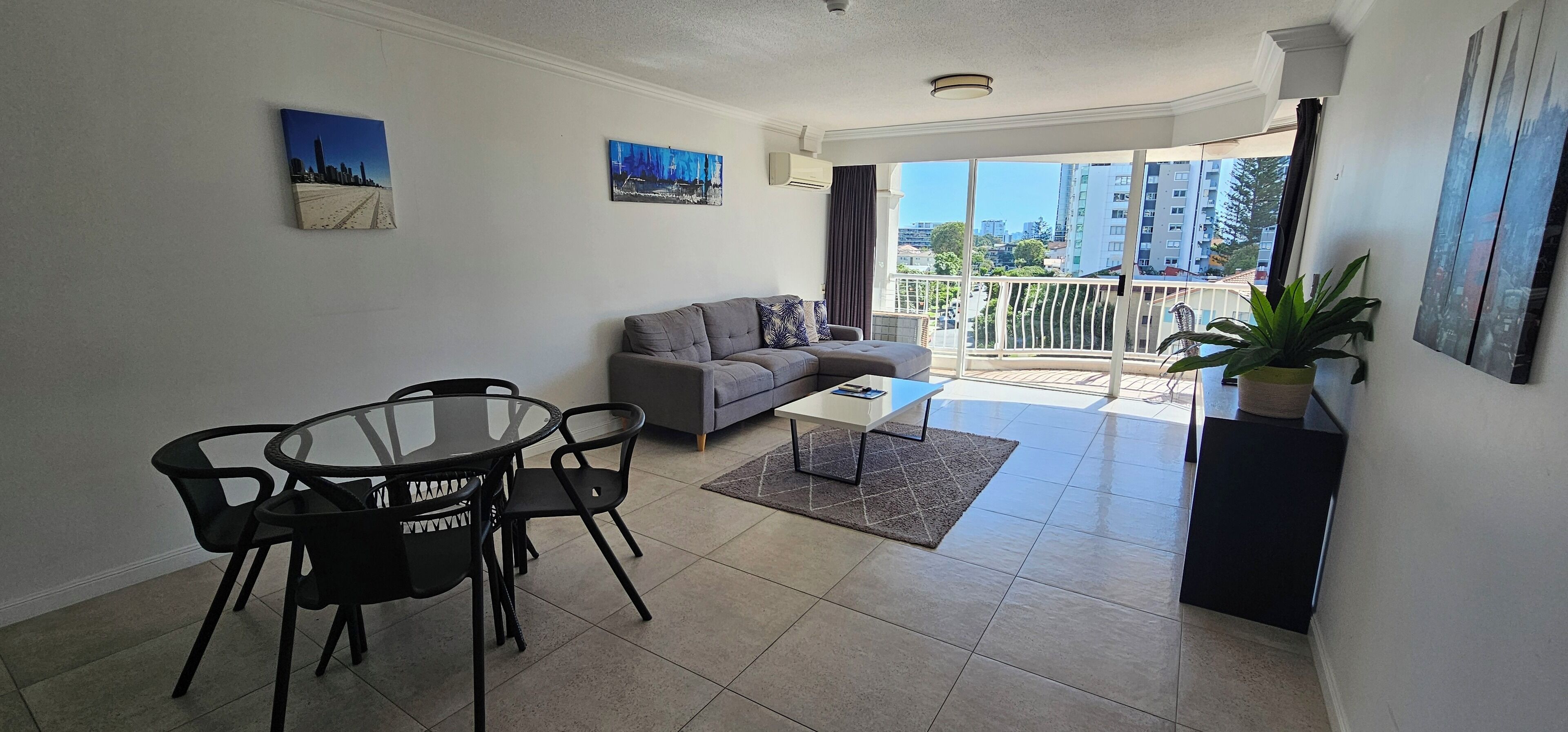 A modern living room with a grey sofa, a glass coffee table, and a dining area with a round table and chairs. There are two paintings on the wall and a balcony with a view of buildings.