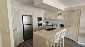 Eine Küche mit weißen Schränken, einem Edelstahlkühlschrank, einem Backofen, einer Mikrowelle und einem Spülbecken. An der Kücheninsel stehen zwei weiße Stühle.