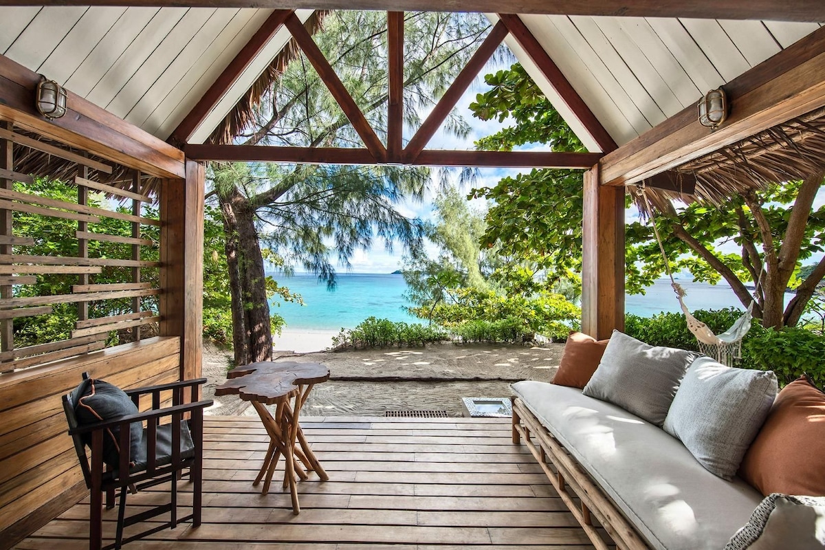 Terraza de madera con sofá y una mesa, con vista a una playa con árboles y una vista despejada del océano.
