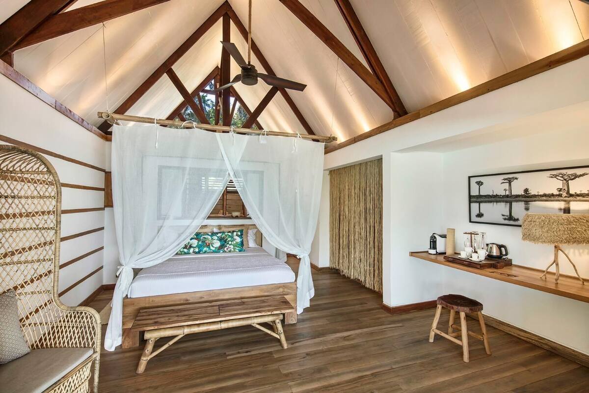 Ein Schlafzimmer mit Holzboden, einem Himmelsbett, einem Korbstuhl, einer Holzbank und einem Regal mit Tee-Service.