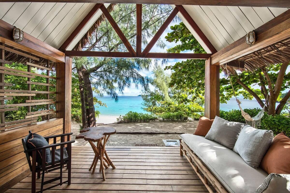 Eine Holzterrasse mit einer Couch und einem Tisch, mit Blick auf einen Strand mit Bäumen und freiem Blick auf den Ozean.