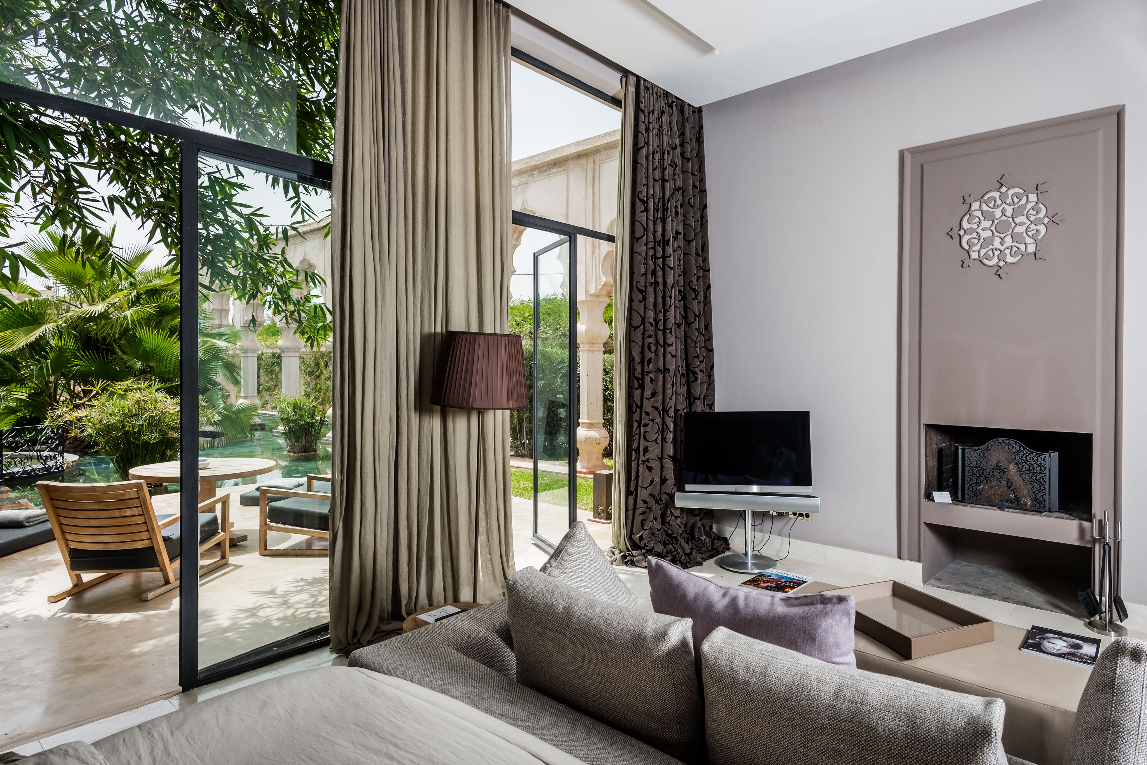 A modern living room with a sofa, TV, and fireplace. There is a glass door leading to a patio with outdoor furniture and plants.
