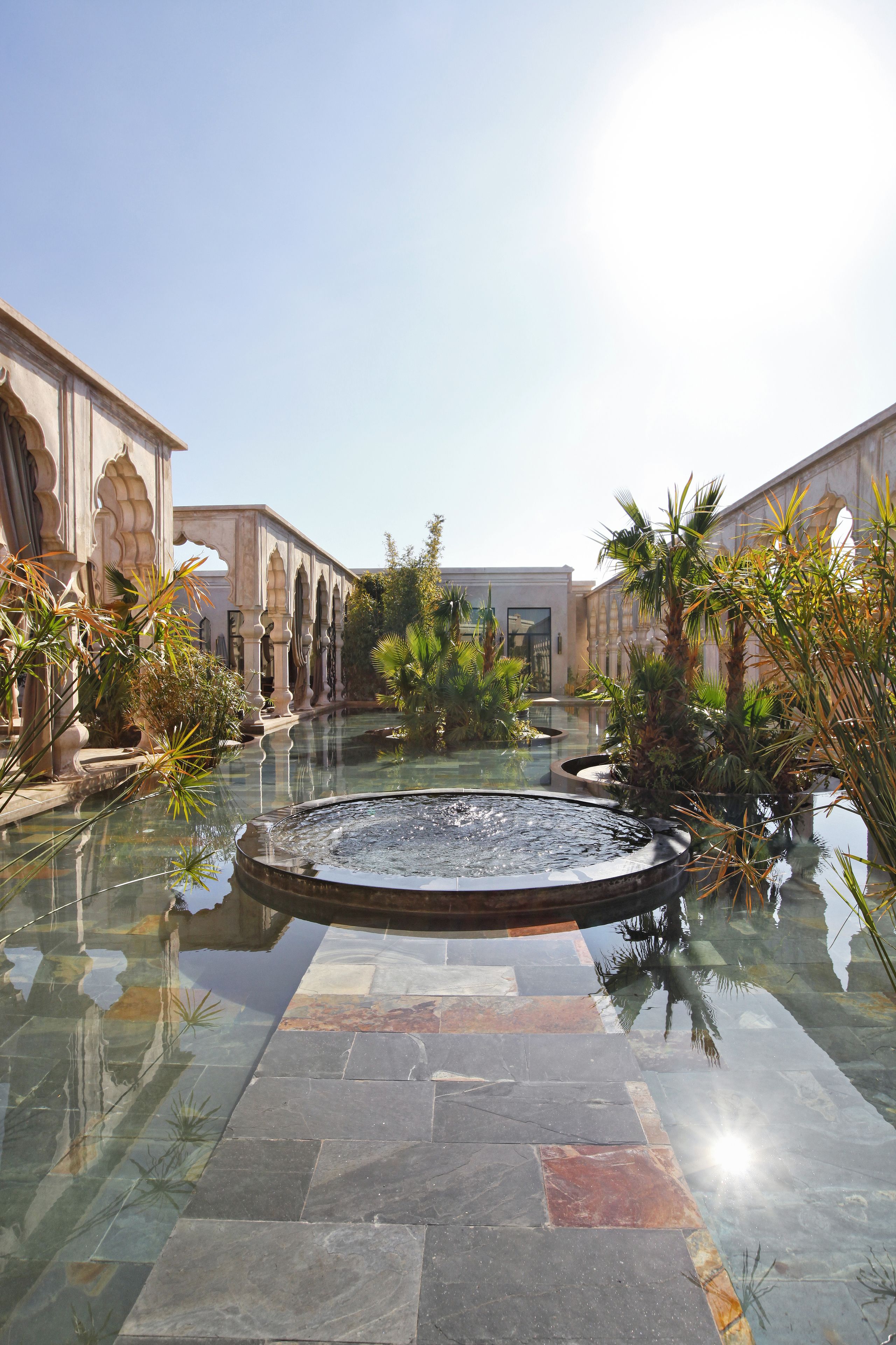 A courtyard with a central water feature, surrounded by palm trees and stone pathways.