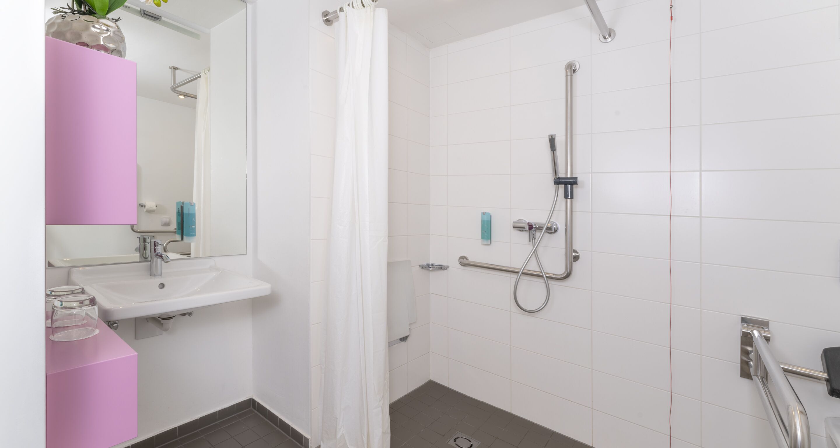 Ein modernes Badezimmer mit einem weißen Waschbecken, einem Spiegel, einer Dusche mit Duschbrause und einer Haltestange.