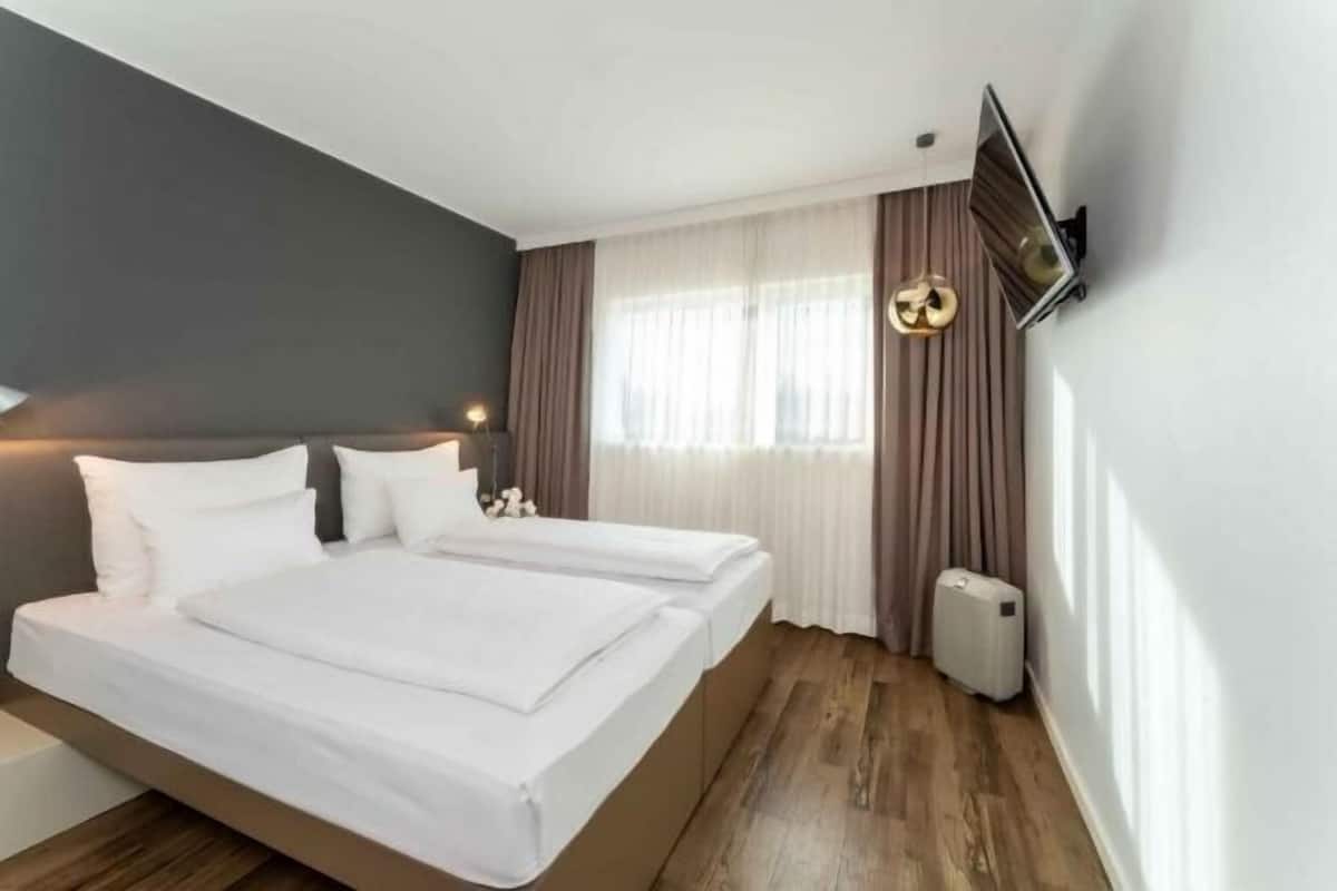 A modern hotel room with a large bed, a flat-screen TV mounted on the wall, and a window with sheer curtains.