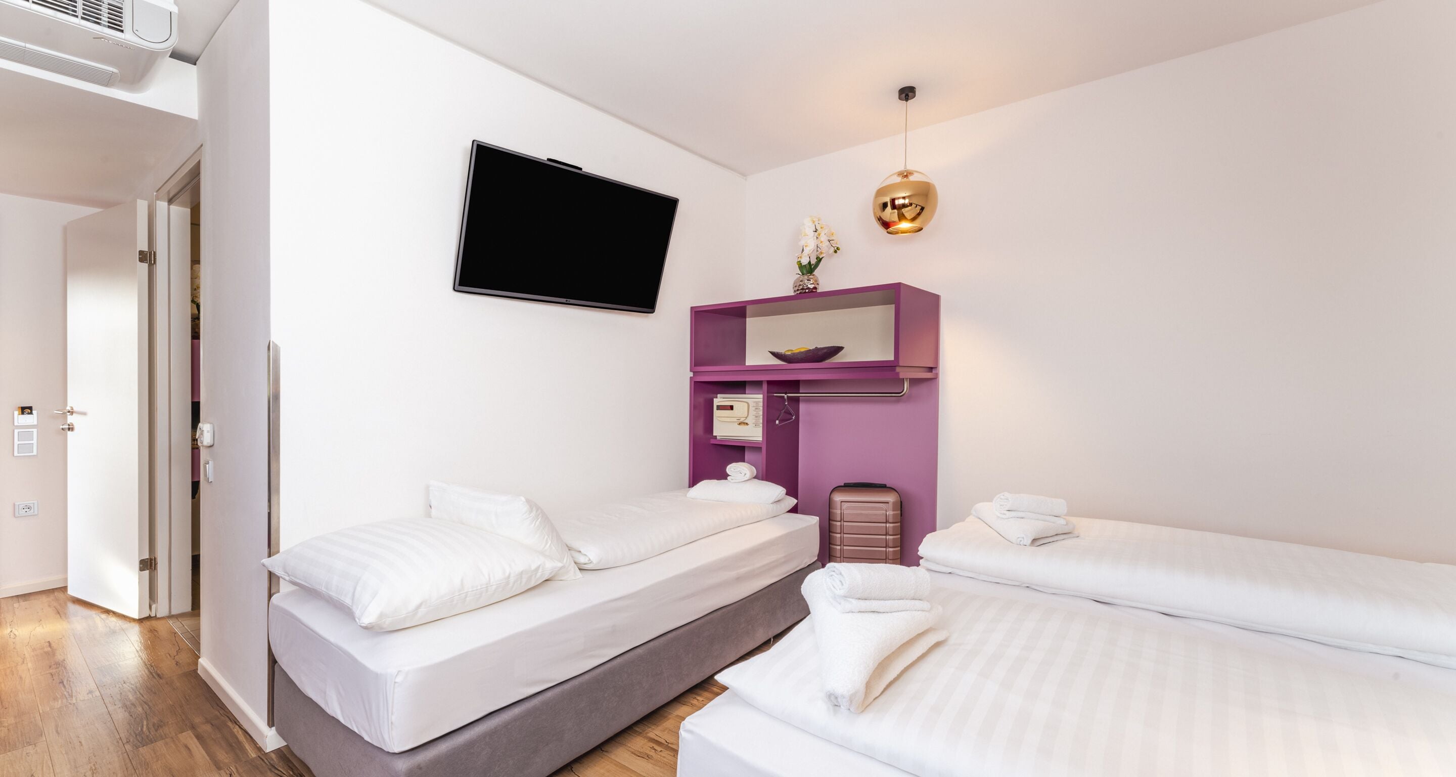 Habitación de hotel con dos camas, un televisor, una estantería morada con un espejo y una mesita con un jarrón de flores.