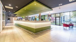 A modern reception area with a green and white counter, a seating area with red chairs, and large windows providing a view of the outdoors. - PLAZA INN Graz Messe (Graz)