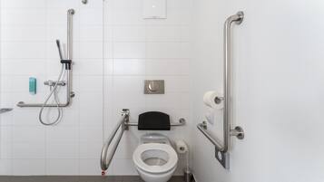 A bathroom with a white tiled wall, a white toilet, a black toilet seat, a handrail, a grab bar, a showerhead, and a soap dispenser.