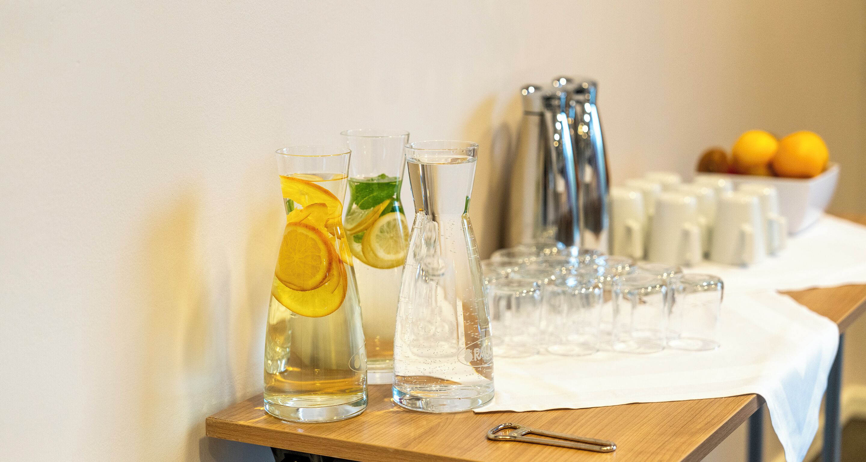 Twee glazen water met citroenpartjes op een houten tafel, een waterfles en een schaal met mandarijnen op de achtergrond.