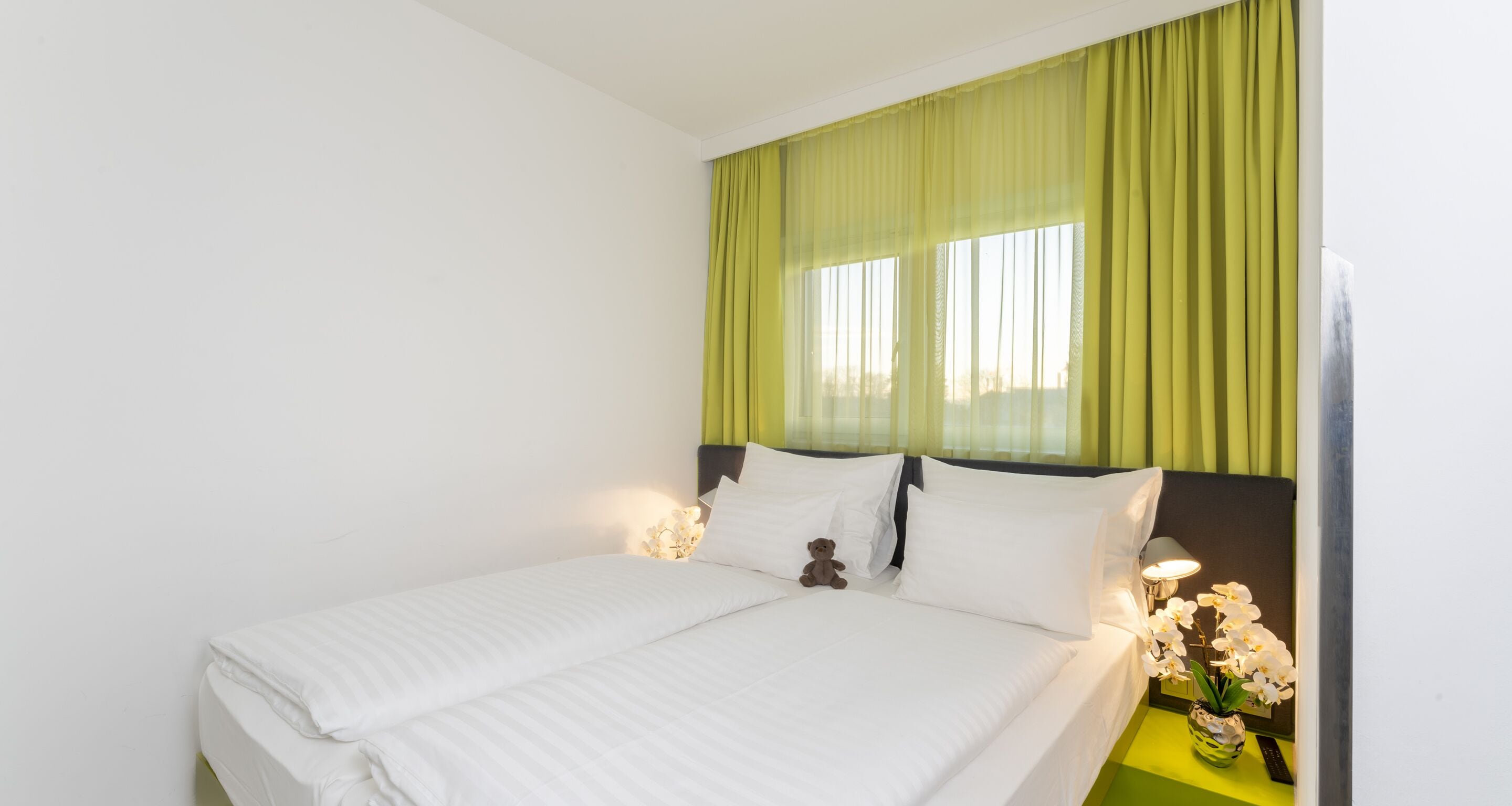 Una cama bien hecha con sábanas blancas, un osito de peluche sobre las almohadas y una mesita de noche con una lámpara y flores.