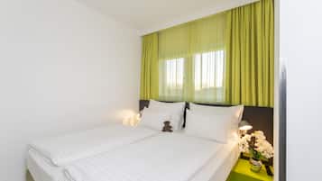 A neatly made bed with white linens, a teddy bear on the pillows, and a nightstand with a lamp and flowers.