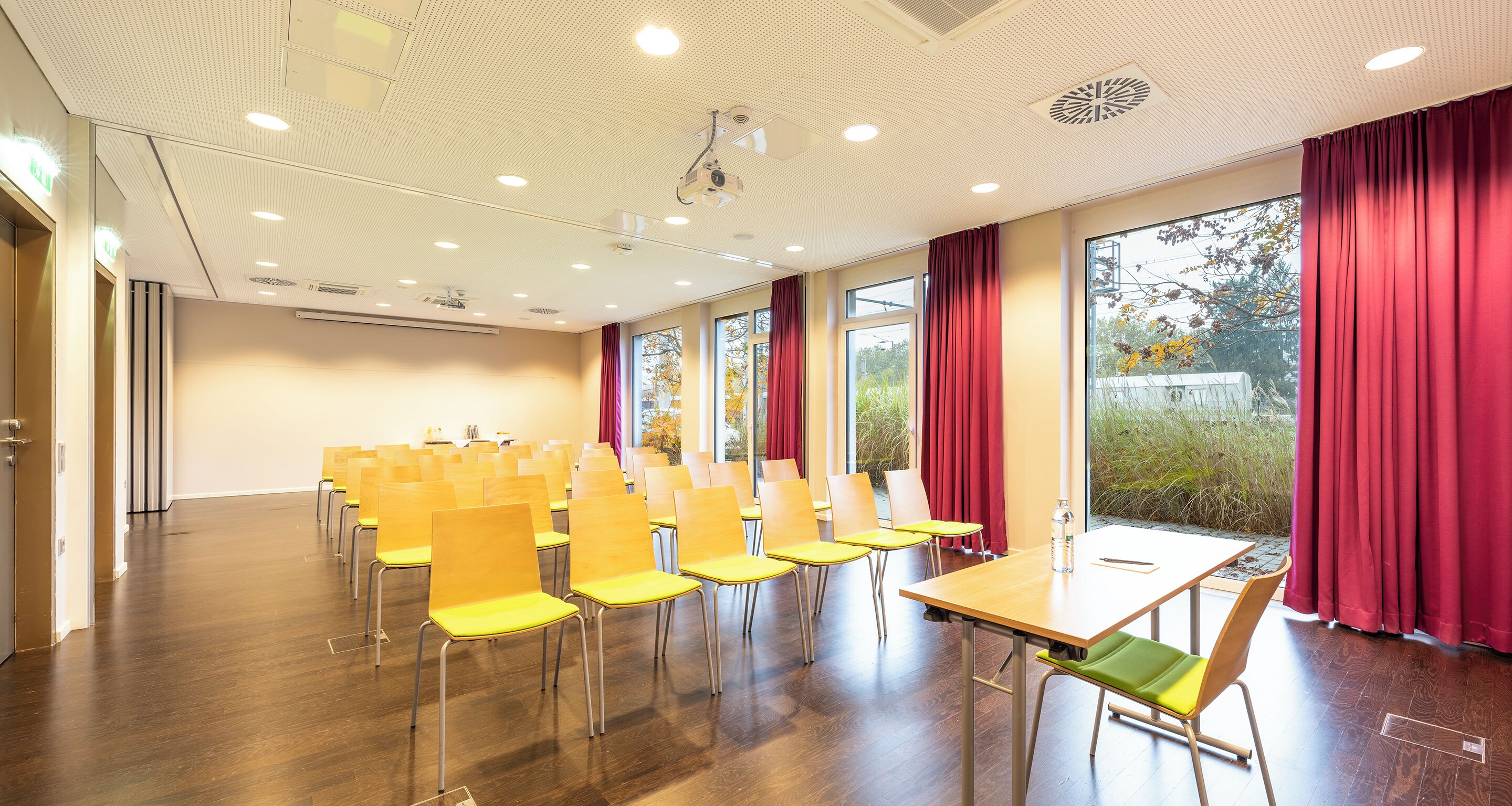 A conference room with rows of yellow chairs, a wooden table, and large windows with red curtains.