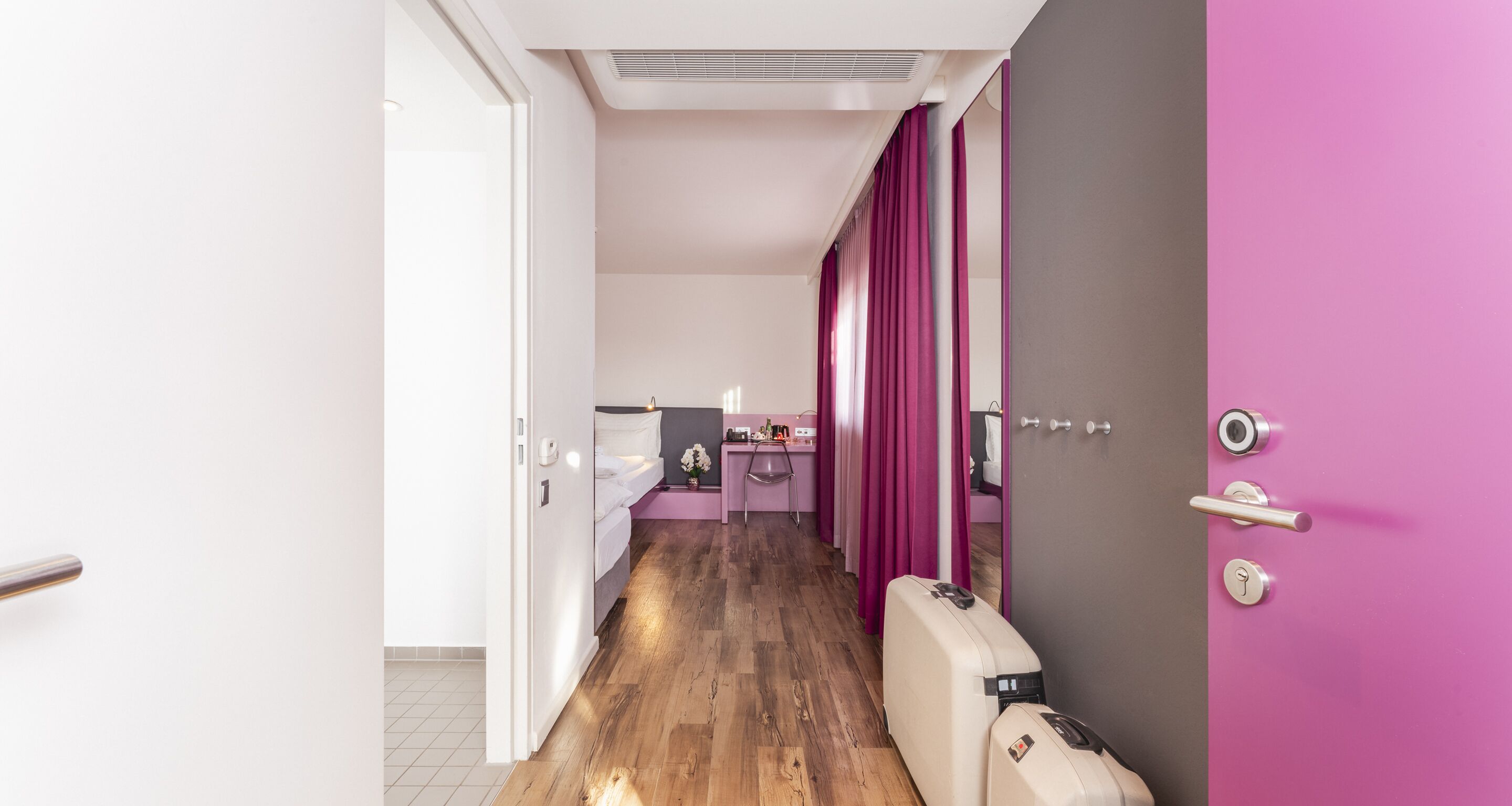 A hotel room with a wooden floor, a bed, a desk with a chair, a pink curtain, and a suitcase.