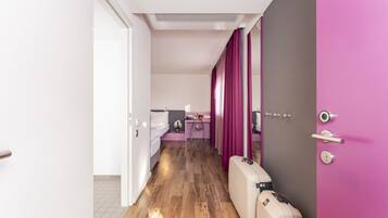 Una habitación de hotel con piso de madera, una cama, un escritorio con silla, una cortina rosa y una maleta.