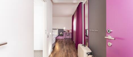 Una habitación de hotel con piso de madera, una cama, un escritorio con silla, una cortina rosa y una maleta.