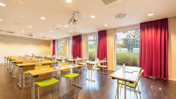 Un salón de conferencias con filas de mesas y sillas, ventanas amplias con cortinas rojas e iluminación empotrada en el techo, además de un proyector.