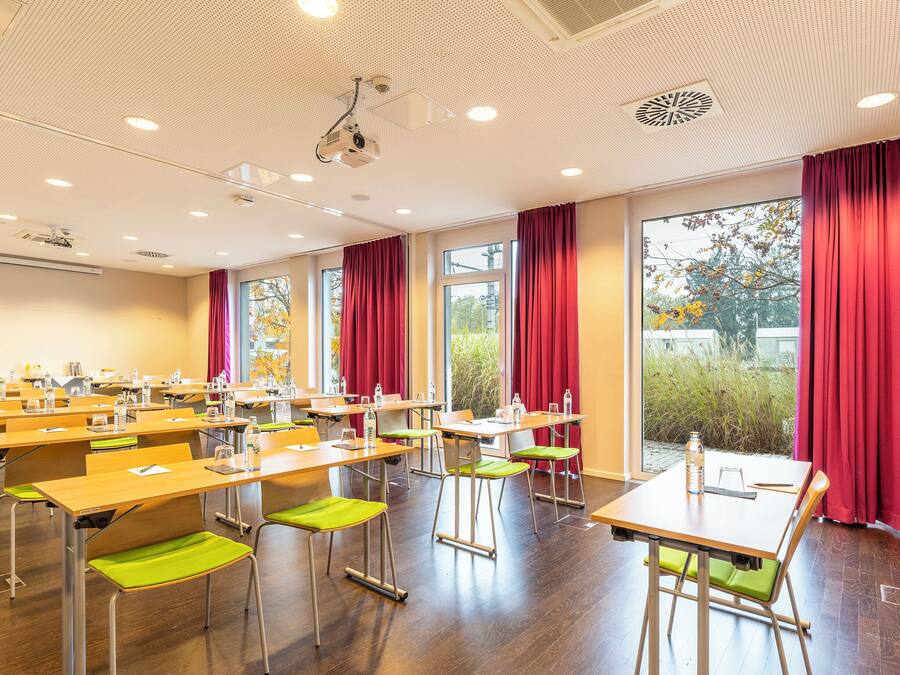 Un salón de conferencias con filas de mesas y sillas, ventanas amplias con cortinas rojas e iluminación empotrada en el techo, además de un proyector.