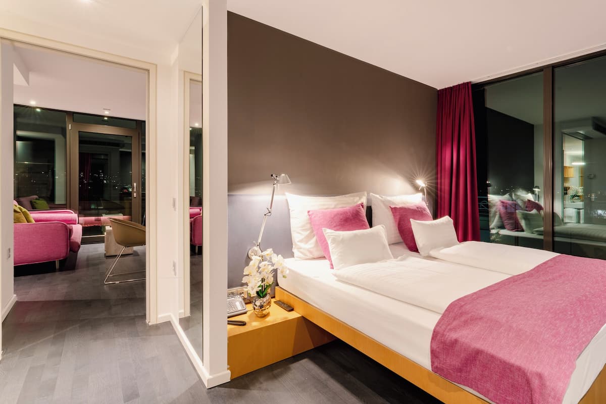 A modern hotel room with a large bed, a nightstand, a desk, and a view of the city through large windows.