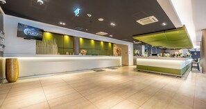 A modern hotel reception area with a flat-screen TV displaying the hotel name, a reception desk, and a green accent wall.