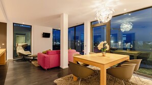A modern living room with a wooden dining table, two pink sofas, and a large window offering a city view. - PLAZA INN Graz Messe (Graz)