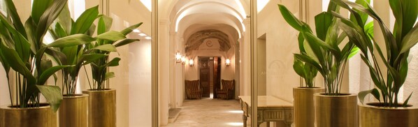 A hallway with large potted plants, a marble floor, and a grand archway leading to a room.