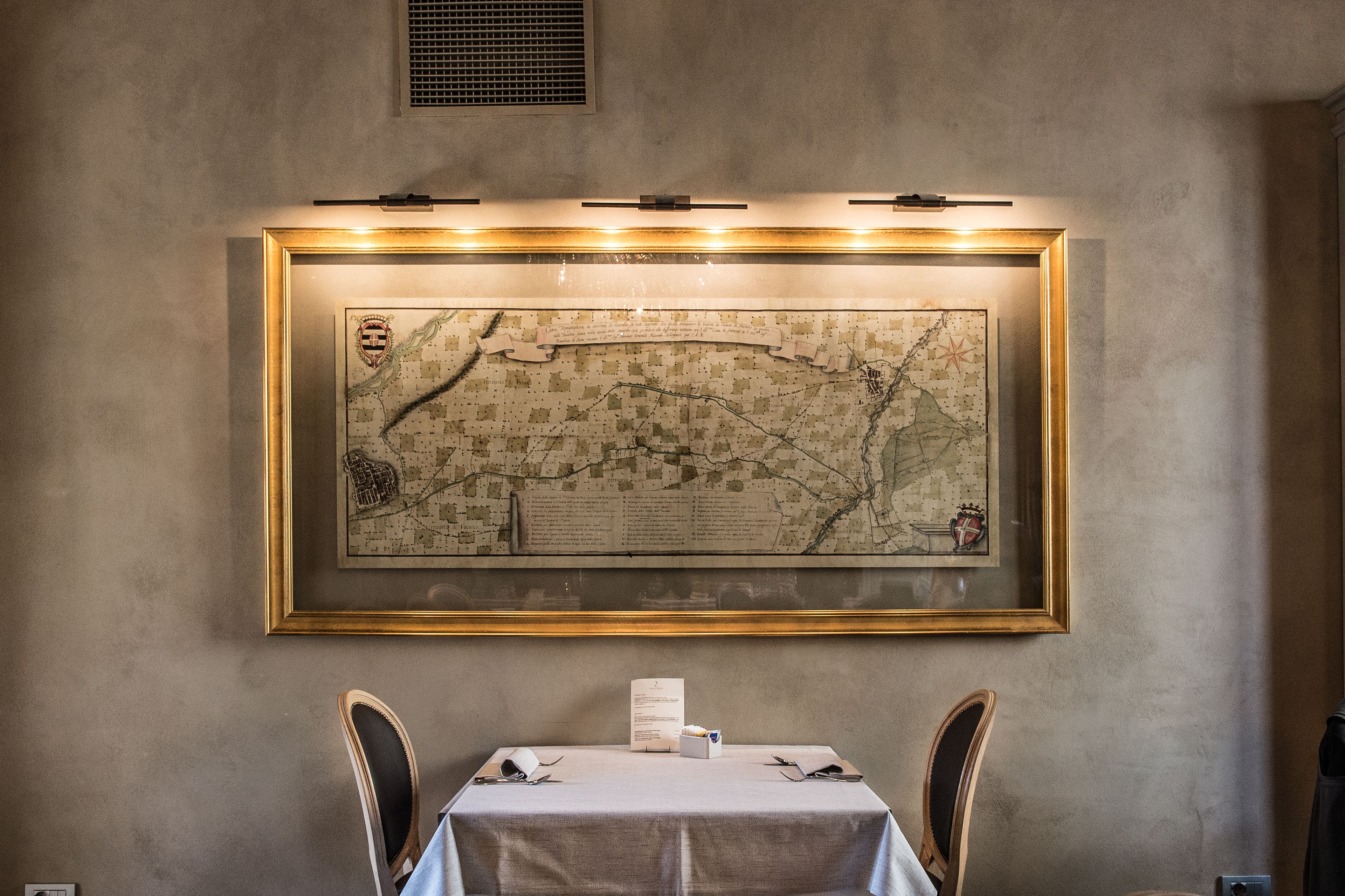 Un coin repas avec une table dressée pour deux personnes, une carte encadrée accrochée au mur et un menu posé sur la table.