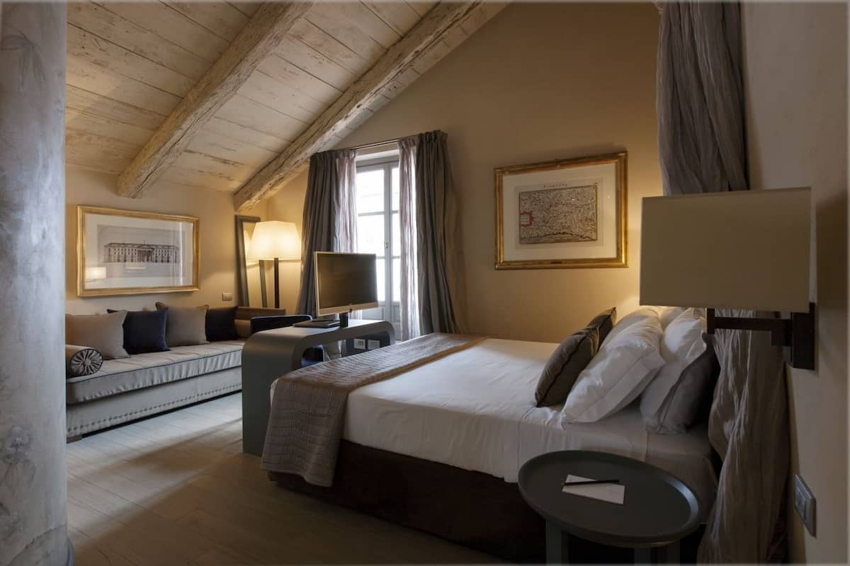 Quarto de hotel com cama grande, sofá, escrivaninha com computador e janela com cortinas.