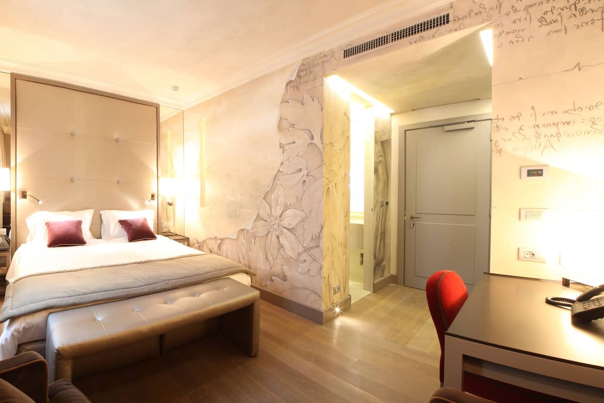 Quarto de hotel com uma cama grande, um banco, uma escrivaninha com telefone e um espelho.