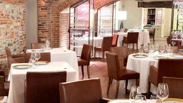 A restaurant with brick walls and white tablecloths, featuring round tables and brown leather chairs.