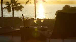 A balcony with a view of the sunset over the ocean, two glasses of white wine, and a bottle of wine on a wicker table.