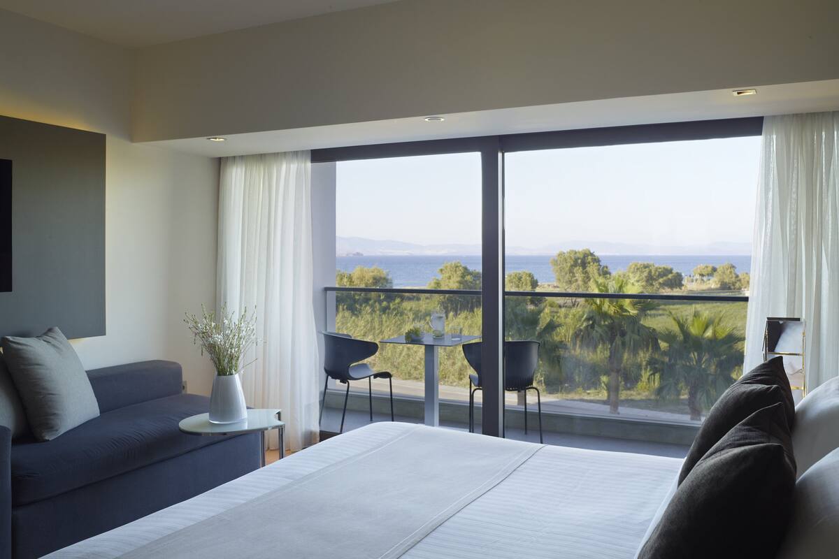 Een moderne slaapkamer met een groot bed, een bank, een klein tafeltje en een balkon met uitzicht op zee en groen.