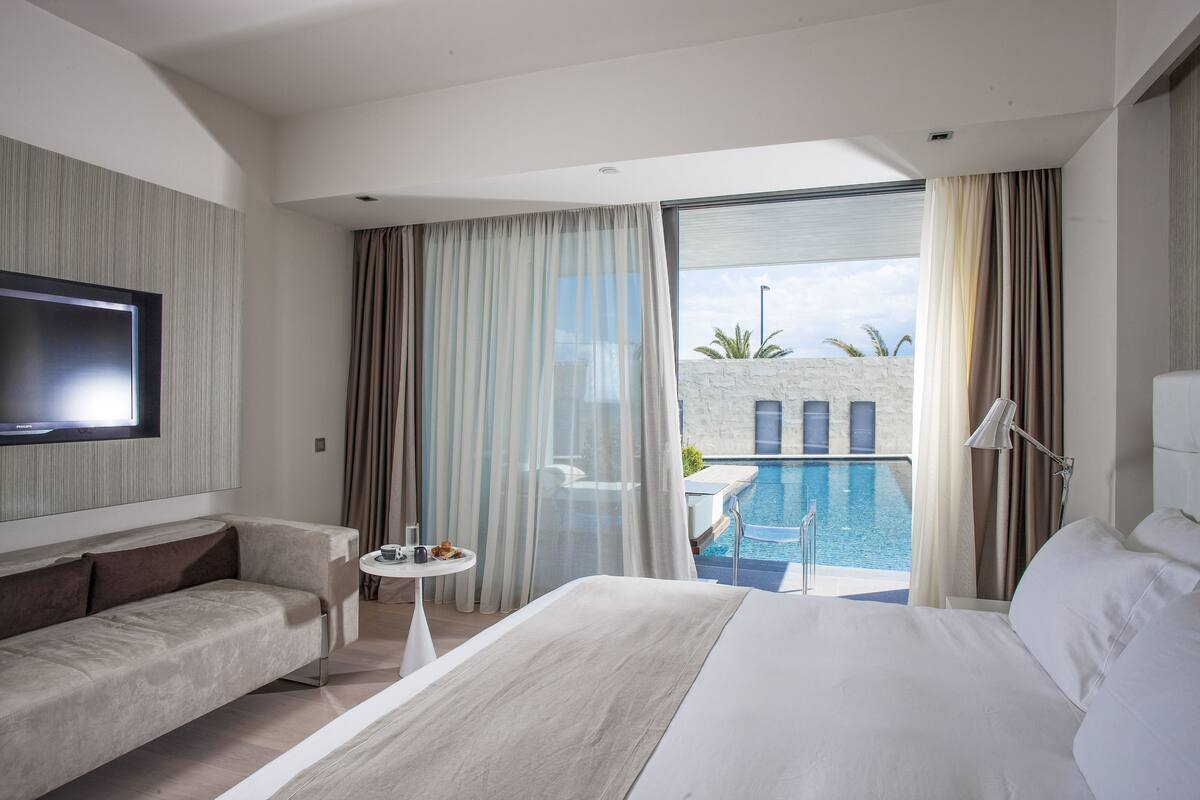 Een moderne hotelkamer met een groot bed, een bank, een aan de muur bevestigde televisie, een klein tafeltje met een drankje en uitzicht op een zwembad.