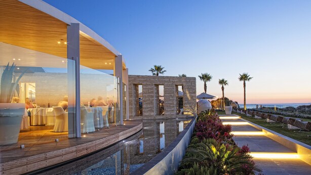 A modern outdoor dining area with a wooden deck, glass walls, and a view of the ocean.