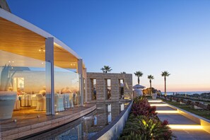 Een moderne eethoek buiten, met een houten terras, glazen wanden en uitzicht op zee.