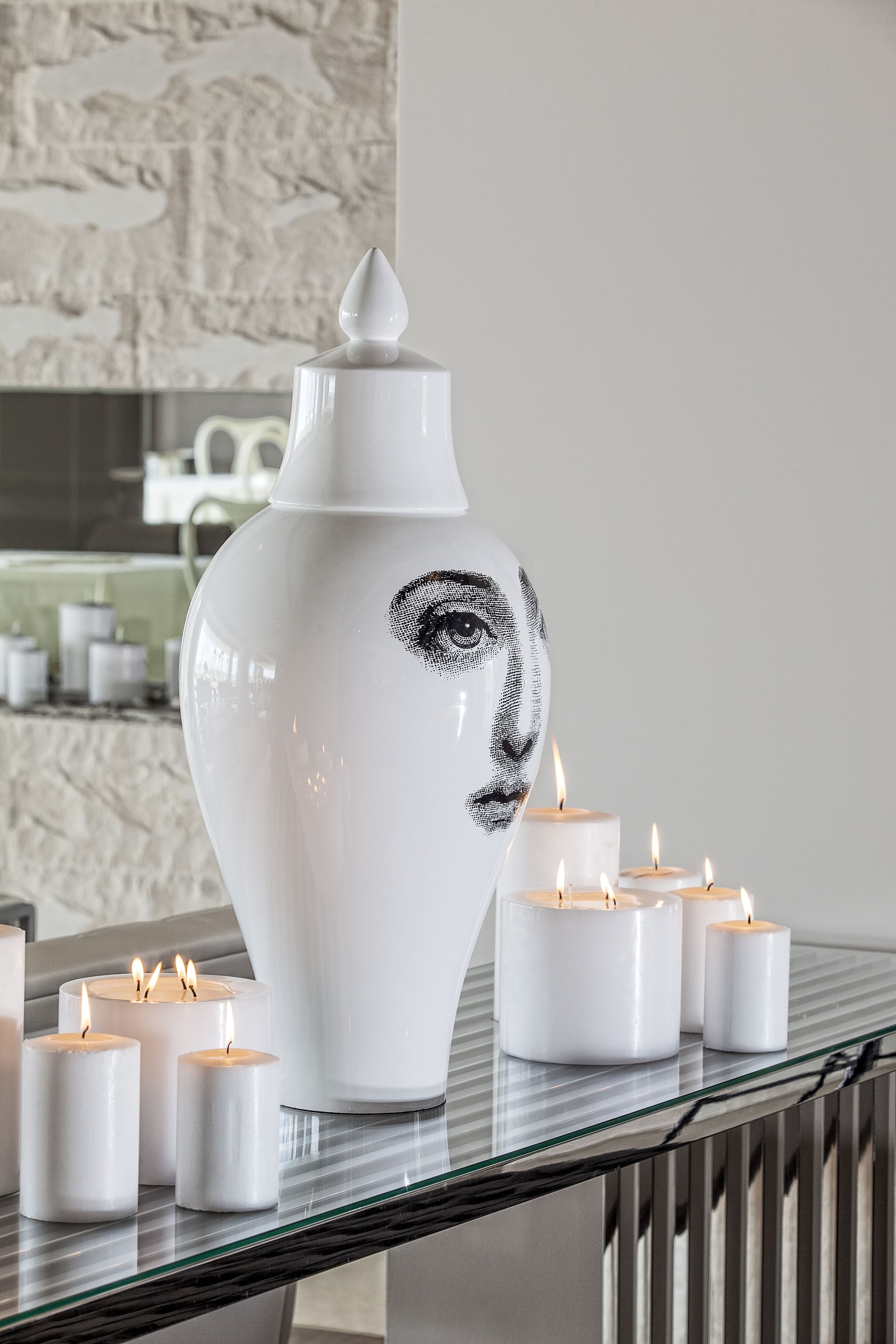 A white vase with a face design on a glass table surrounded by lit candles.